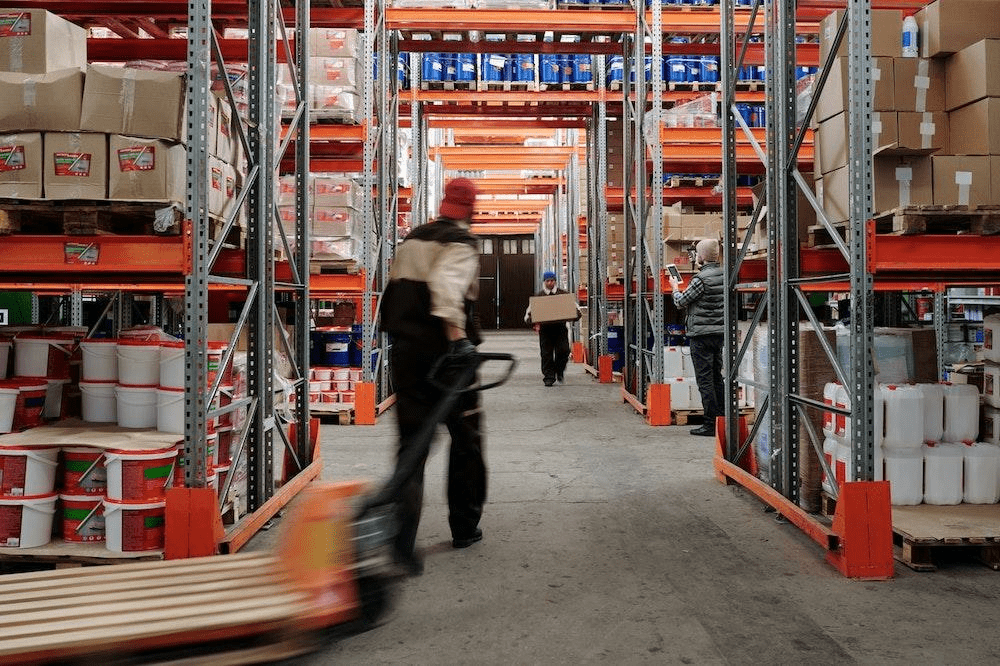 in a warehouse, one worker with a pallet jack, another carrying a box, another checking inventory counts